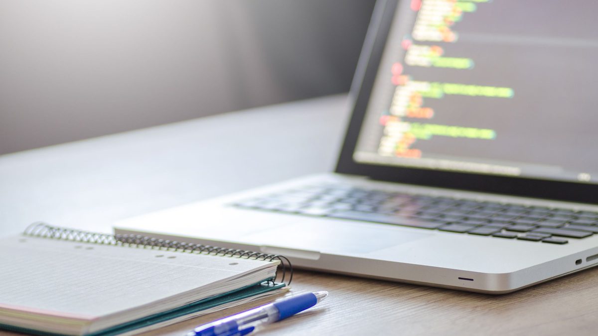 A laptop showing lines of code beside a notebook and pen.